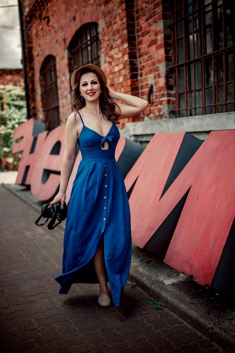 Kobieta w niebieskiej sukience i kapeluszu pozuje z lustrzanką na tle ceglanej ściany z dużymi, czerwono-czarnymi literami.