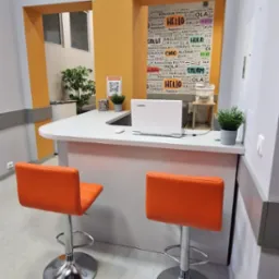 Recepcja szkoły językowej z logo 'Dobra Szkoła', plakat z powitaniami w różnych językach, laptop Lenovo, dwie pomarańczowe krzesła barowe i dekoracyjne rośliny w doniczkach.