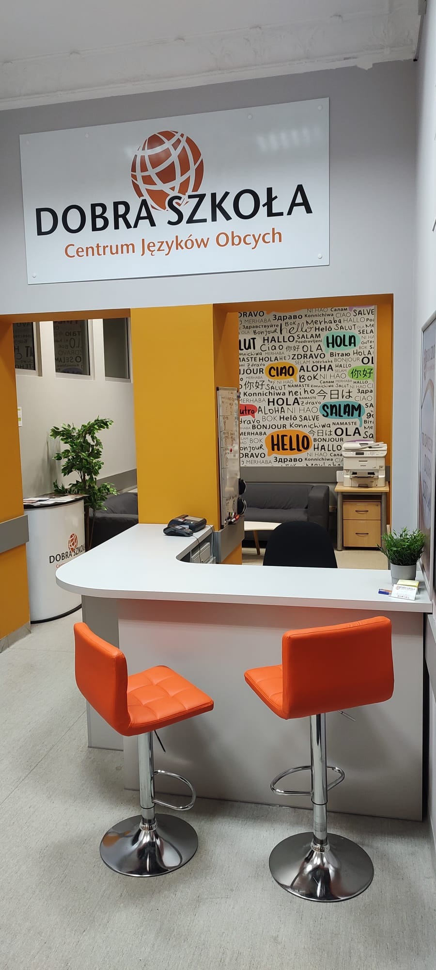 Recepcja szkoły językowej z pomarańczowymi krzesłami barowymi, widoczny napis 'Dobra Szkoła Centrum Języków Obcych' i ściana z napisem 'Hello' w różnych językach.