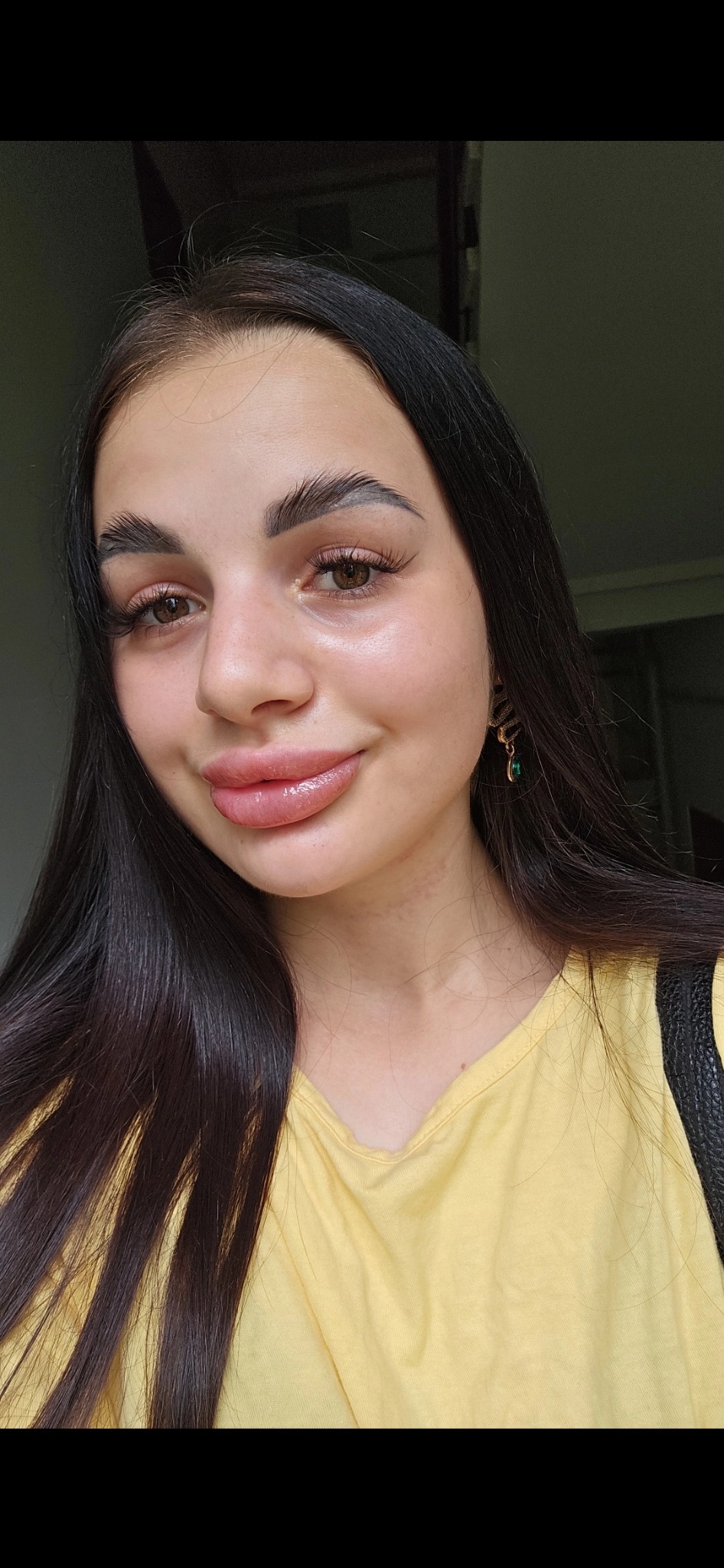 Portret uśmiechniętej kobiety z długimi, ciemnymi włosami, mocno podkreślonymi brwiami i błyszczącymi ustami, ubrana w żółtą koszulkę.