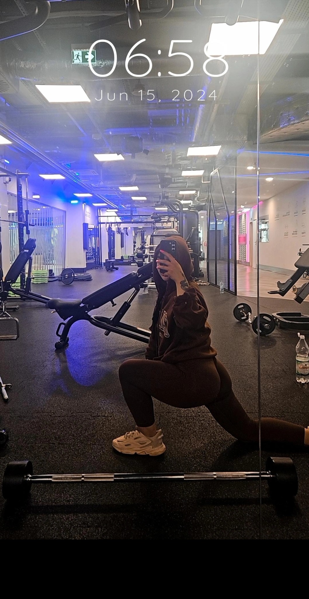 Osoba w brązowym dresie wykonuje wypad w przód w siłowni, widoczne sztangi, ławka do ćwiczeń i lustro, data na ekranie telefonu: 15 czerwca 2024.