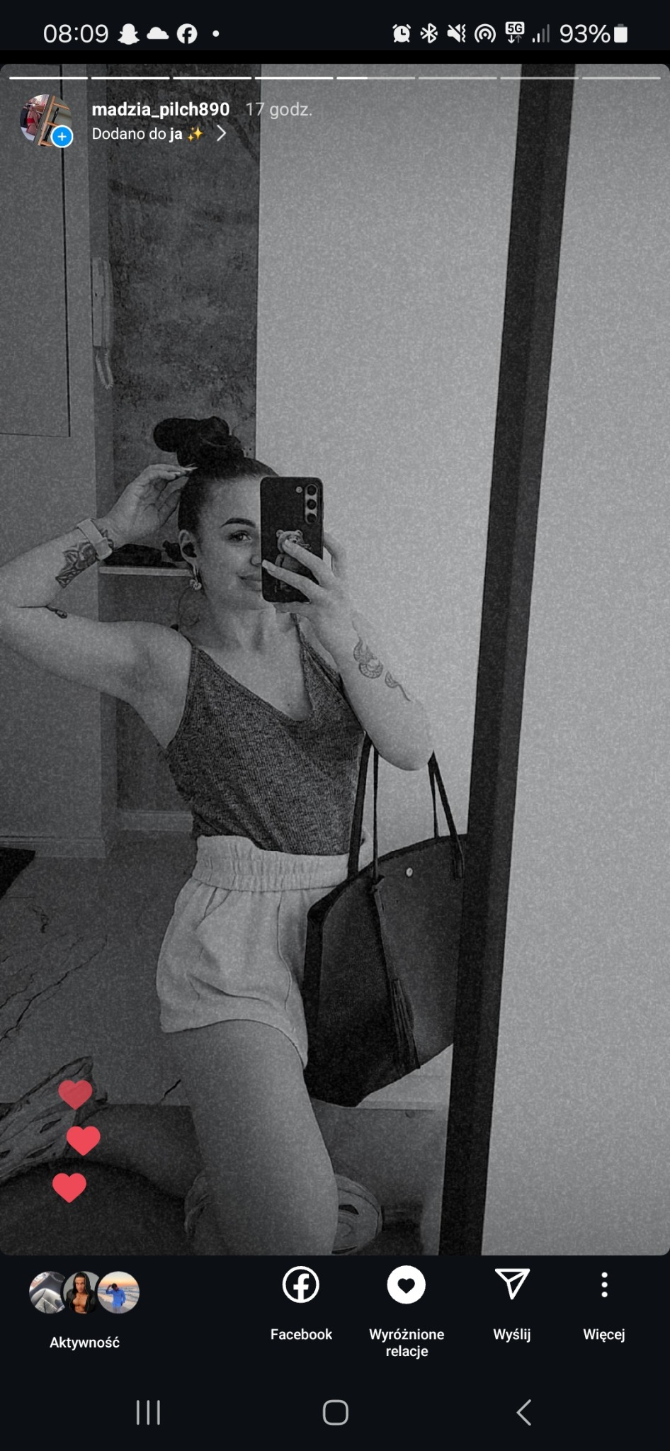 Czarno-białe selfie kobiety w sportowym stroju, z tatuażami na ramionach, trzymającej telefon z naklejką misia, z torbą na ramieniu, stojącej przed lustrem.