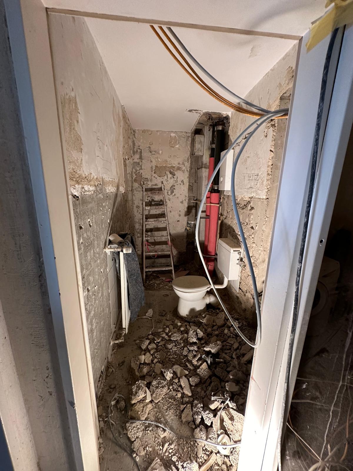 Łazienka w trakcie demontażu: widoczne gruz, odsłonięte instalacje, drabina oparta o ścianę z resztkami starej powłoki, toaleta częściowo zamontowana, przewody elektryczne pod sufitem.