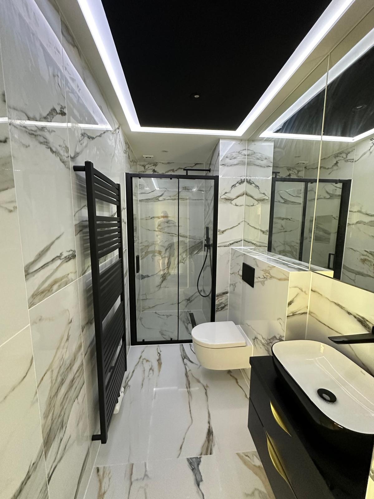 Nowoczesna łazienka z marmurowymi płytkami, czarną kabiną prysznicową, grzejnikiem drabinkowym i umywalką nablatową na czarnej szafce z złotymi uchwytami, oświetlenie LED w suficie.