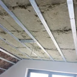 Sufit podwieszany w trakcie montażu: widoczna izolacja z wełny mineralnej między metalowymi profilami, częściowo ukończona konstrukcja w poddaszu z oknem.