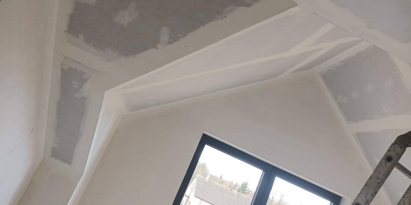 Wnętrze pokoju w trakcie szpachlowania sufitu i ścian, widoczne okno z czarną ramą i fragment drabiny.