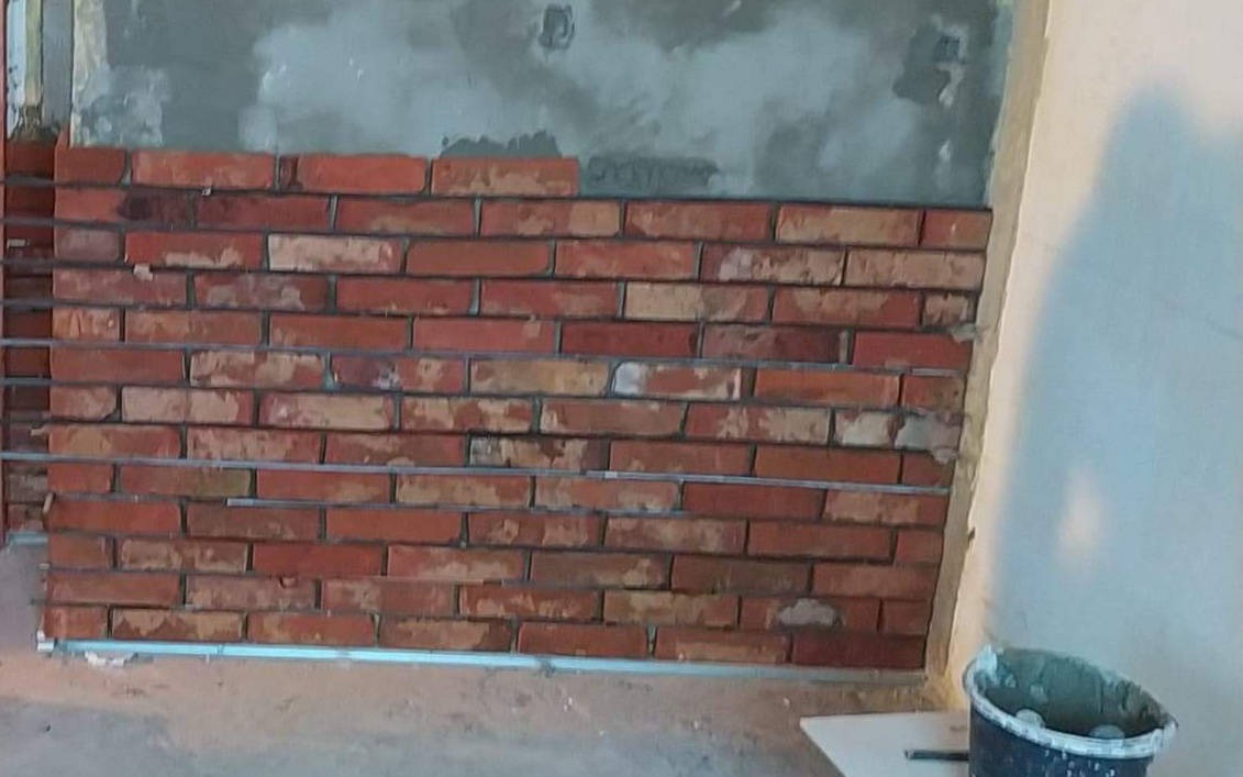 Montaż dekoracyjnej ściany z cegieł wewnątrz pomieszczenia, widoczne metalowe profile i wiadro z zaprawą.