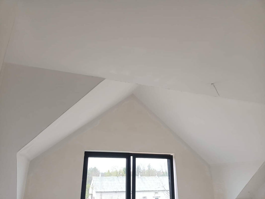 Poddasze z oknem po szpachlowaniu, widoczne niedoskonałości na łączeniach płyt kartonowo-gipsowych na suficie i ścianach szczytowych.