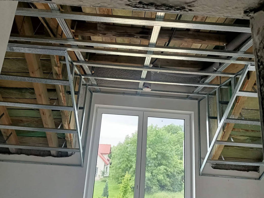 Stelaż z profili metalowych przygotowany do montażu sufitu podwieszanego, widoczny fragment drewnianej konstrukcji dachu i okno dachowe z widokiem na zieleń.