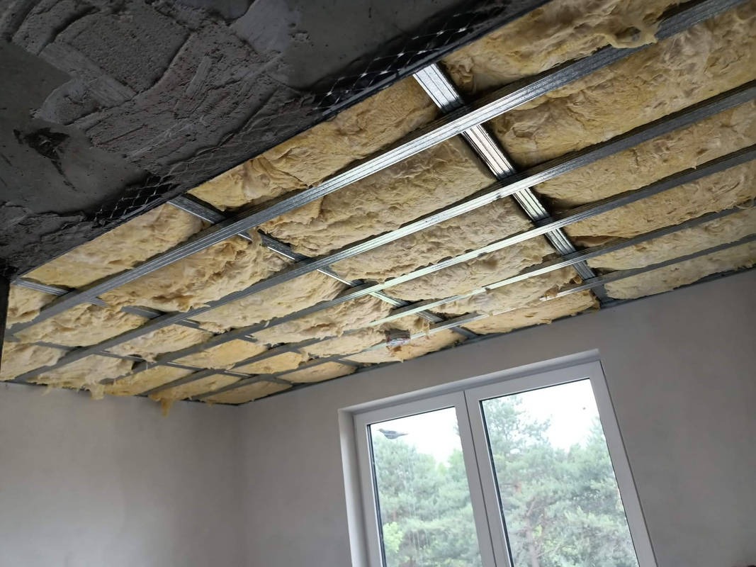 Montaż izolacji z wełny mineralnej w metalowym ruszcie sufitu podwieszanego, widoczne fragmenty surowego stropu i okna z widokiem na drzewa.