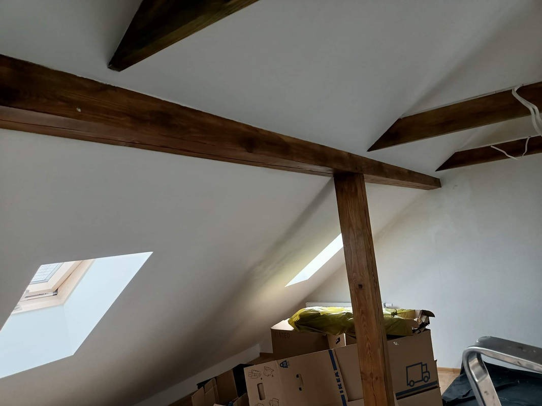 Poddasze po remoncie z widocznymi belkami stropowymi pomalowanymi na ciemny brąz, białe ściany, okna dachowe i kartony Ikea.
