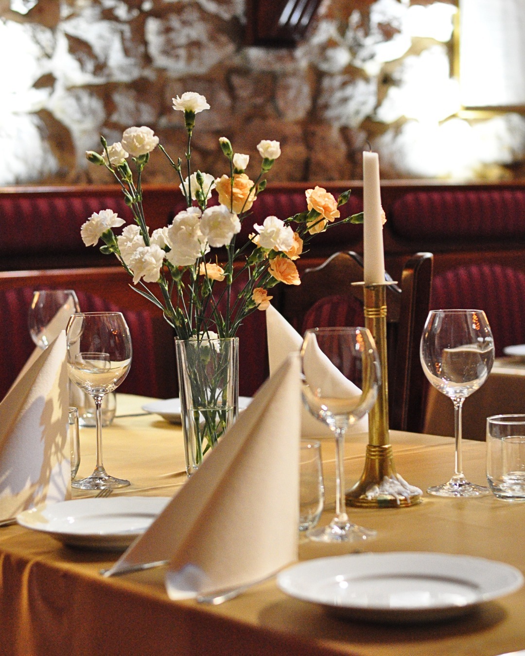 Elegancko nakryty stół z bukietem białych i pomarańczowych goździków w szklanym wazonie, świecą w złotym świeczniku, kieliszkami i złożonymi serwetkami, ustawiony na tle kamiennej ściany...