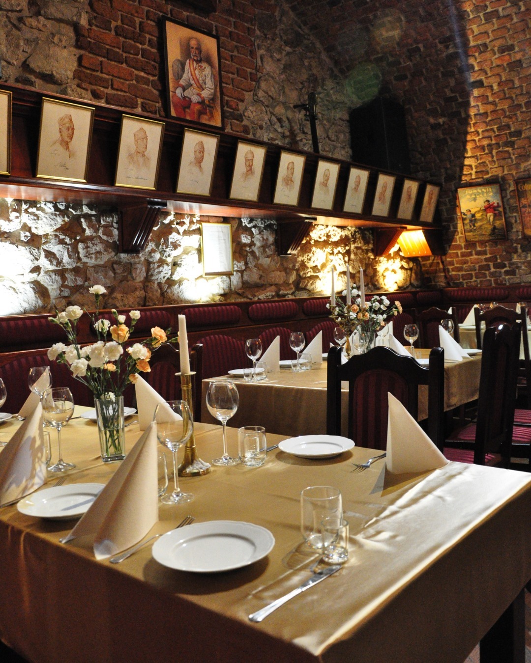 Uroczysta aranżacja stołów w restauracji z ceglanymi ścianami, portretami w ramach i złotymi obrusami, przygotowana na specjalne wydarzenie.