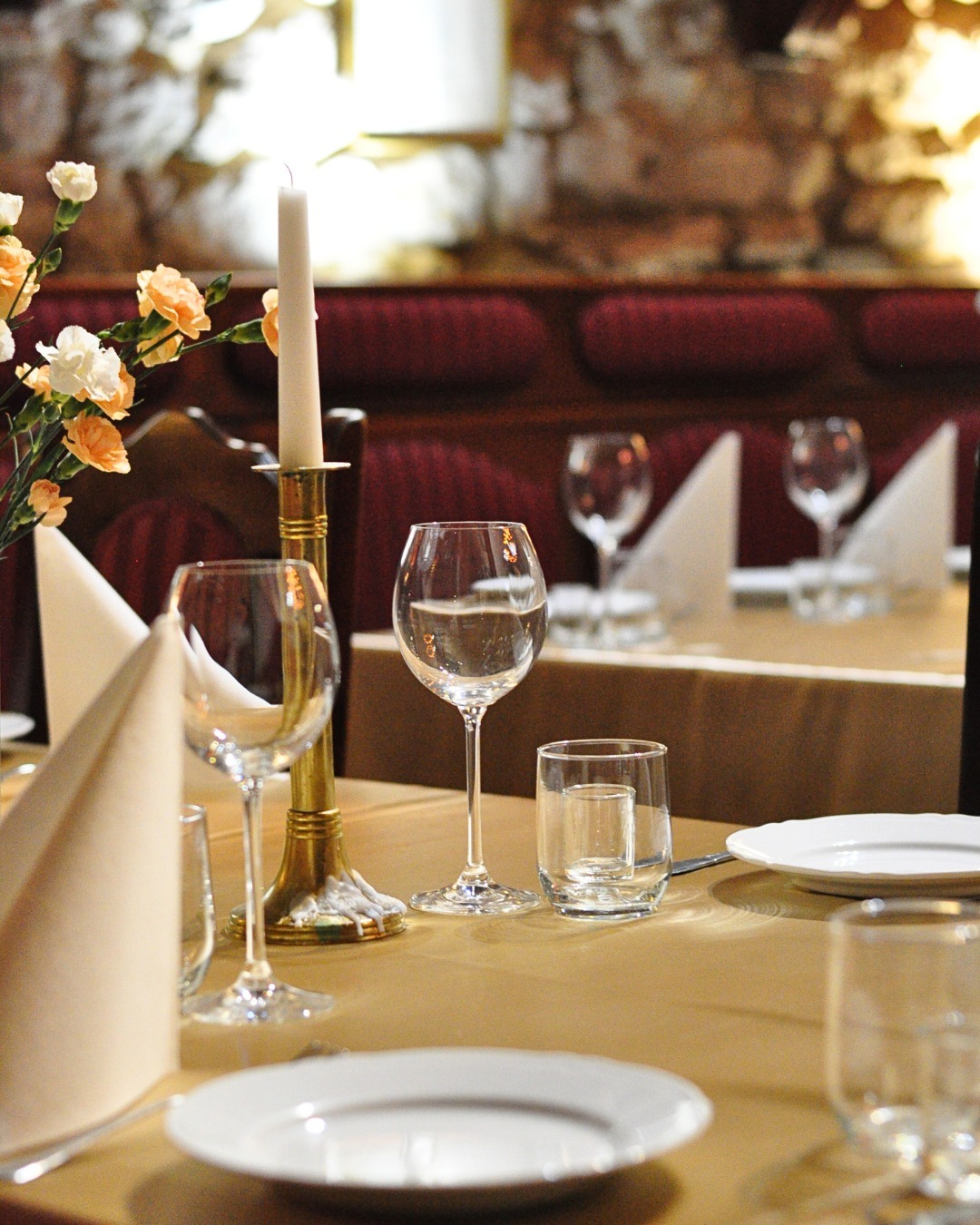 Elegancko nakryty stół z białymi talerzami, kieliszkami, świecą w złotym świeczniku i kompozycją kwiatową w restauracji o ceglanej ścianie w tle.