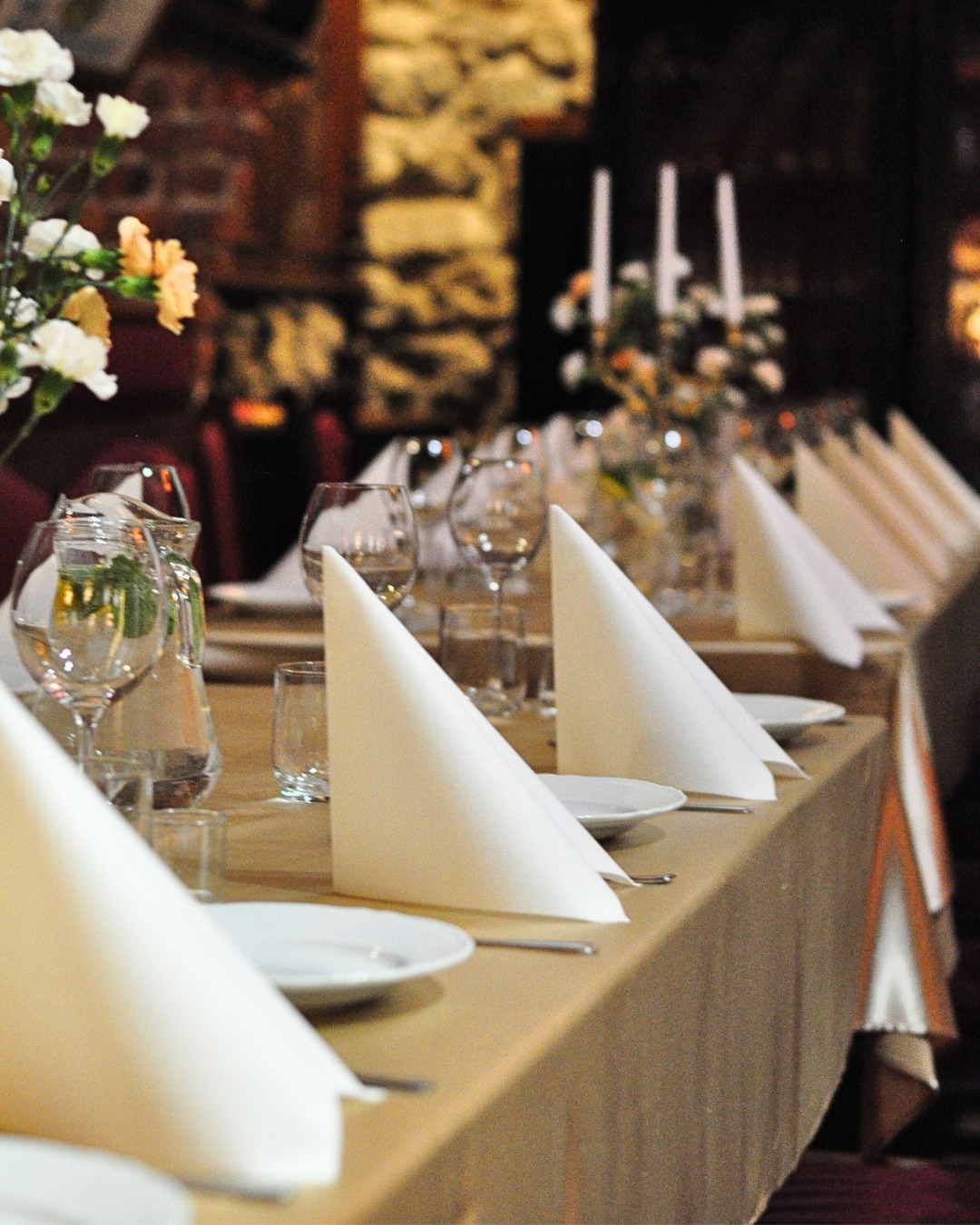 Uroczysty stół z dekoracjami kwiatowymi, nakryty białymi serwetkami złożonymi w stożek, szklanki i zastawa, w tle rustykalna ściana z kamienia.