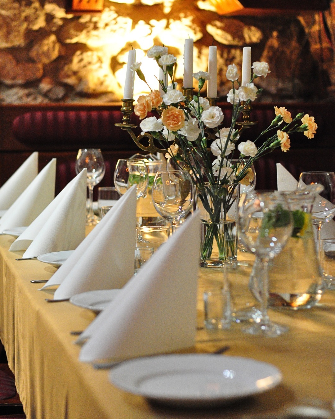 Elegancko nakryty stół z białymi serwetkami ułożonymi w trójkąty, świecznikiem ze świecami i wazonem z białymi i brzoskwiniowymi kwiatami, na tle ściany z kamienia.