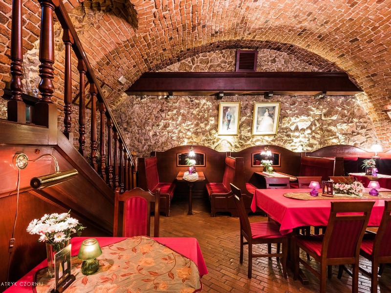 Uroczysta aranżacja wnętrza restauracji z ceglanym sklepieniem, drewnianymi schodami i czerwonymi obrusami, przygotowana na wesele, z portretami pary młodej na ścianie.