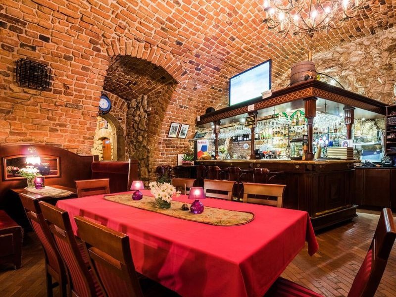 Wnętrze restauracji z ceglanymi ścianami, długim stołem nakrytym czerwonym obrusem, barem z podświetlonymi butelkami i żyrandolem w stylu retro.