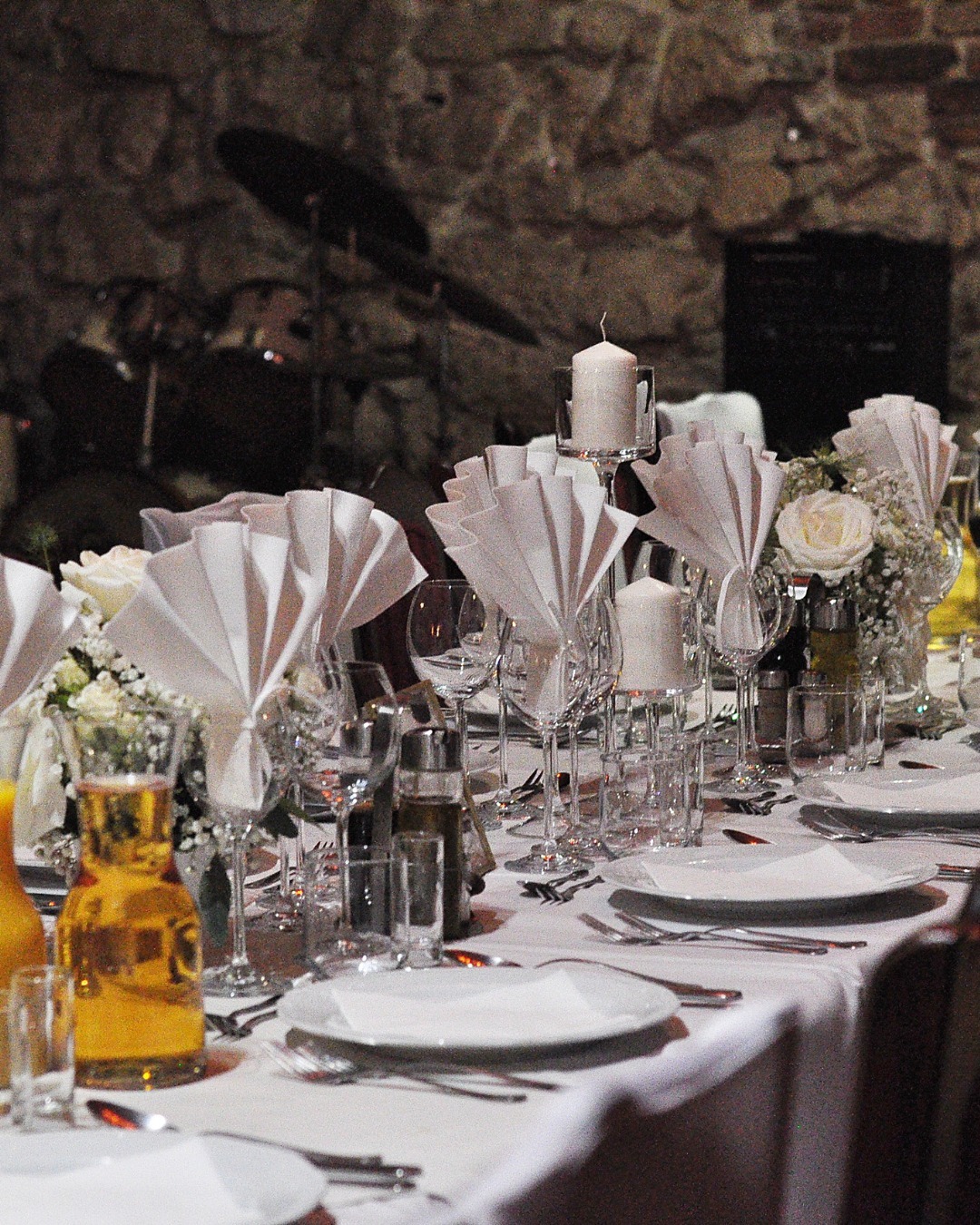 Uroczysty stół z białym obrusem, elegancko złożonymi serwetkami, świecami w szklanych świecznikach, bukietami białych róż i kompletem szkła, w tle widoczny zestaw perkusyjny.