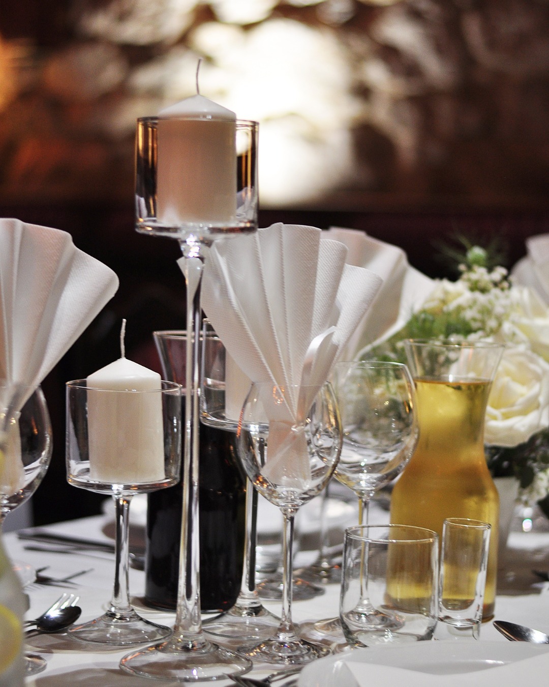 Elegancko nakryty stół z białymi świecami w szklanych świecznikach, kieliszkami, karafką z napojem i dekoracyjnie złożonymi serwetkami, tworzący wyrafinowaną aranżację.