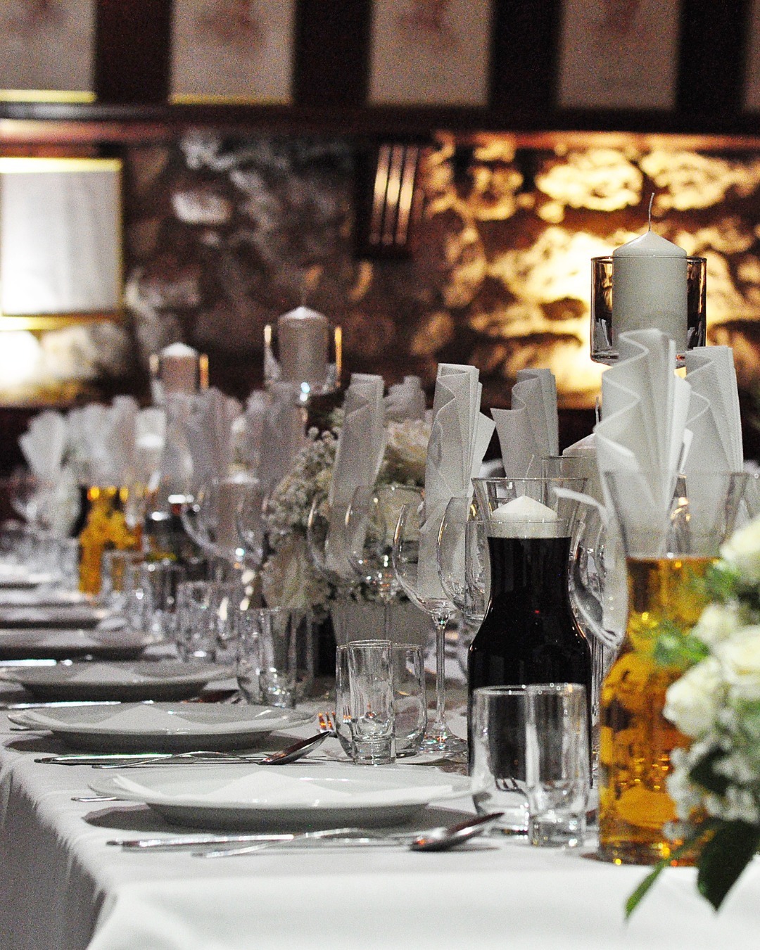 Uroczysty stół nakryty białym obrusem, z elegancką zastawą, kieliszkami, świecami w szklanych świecznikach i kwiatowymi dekoracjami.