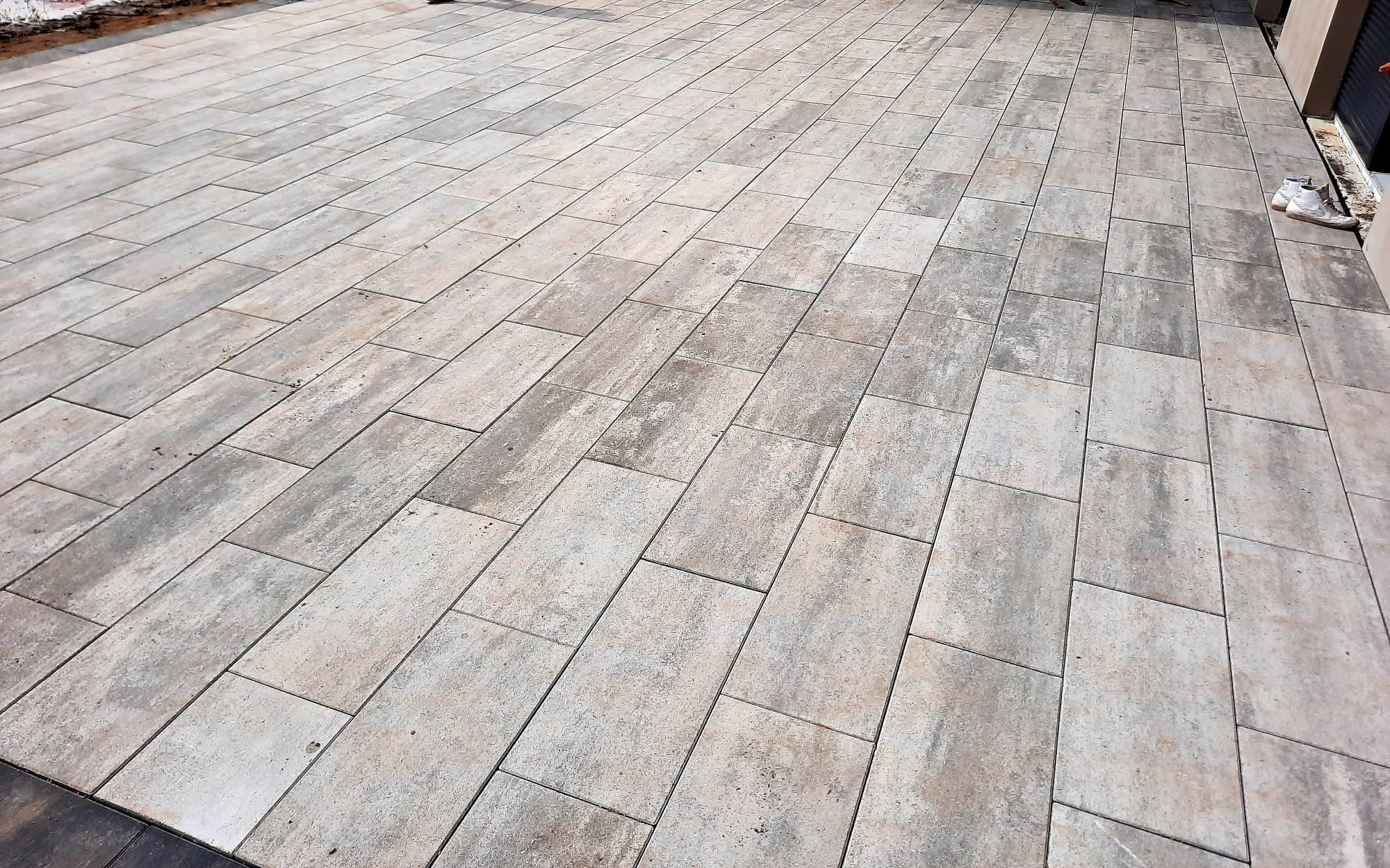 Ułożona nawierzchnia z prostokątnej kostki brukowej w odcieniach beżu i szarości, widoczne białe buty przy ścianie budynku w prawym górnym rogu.
