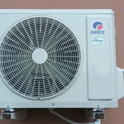 Zewnętrzna jednostka klimatyzacji marki Gree zamontowana na ścianie budynku o różowej elewacji. Widoczne logo i oznaczenie Inverter. Białe przewody i wsporniki montażowe.