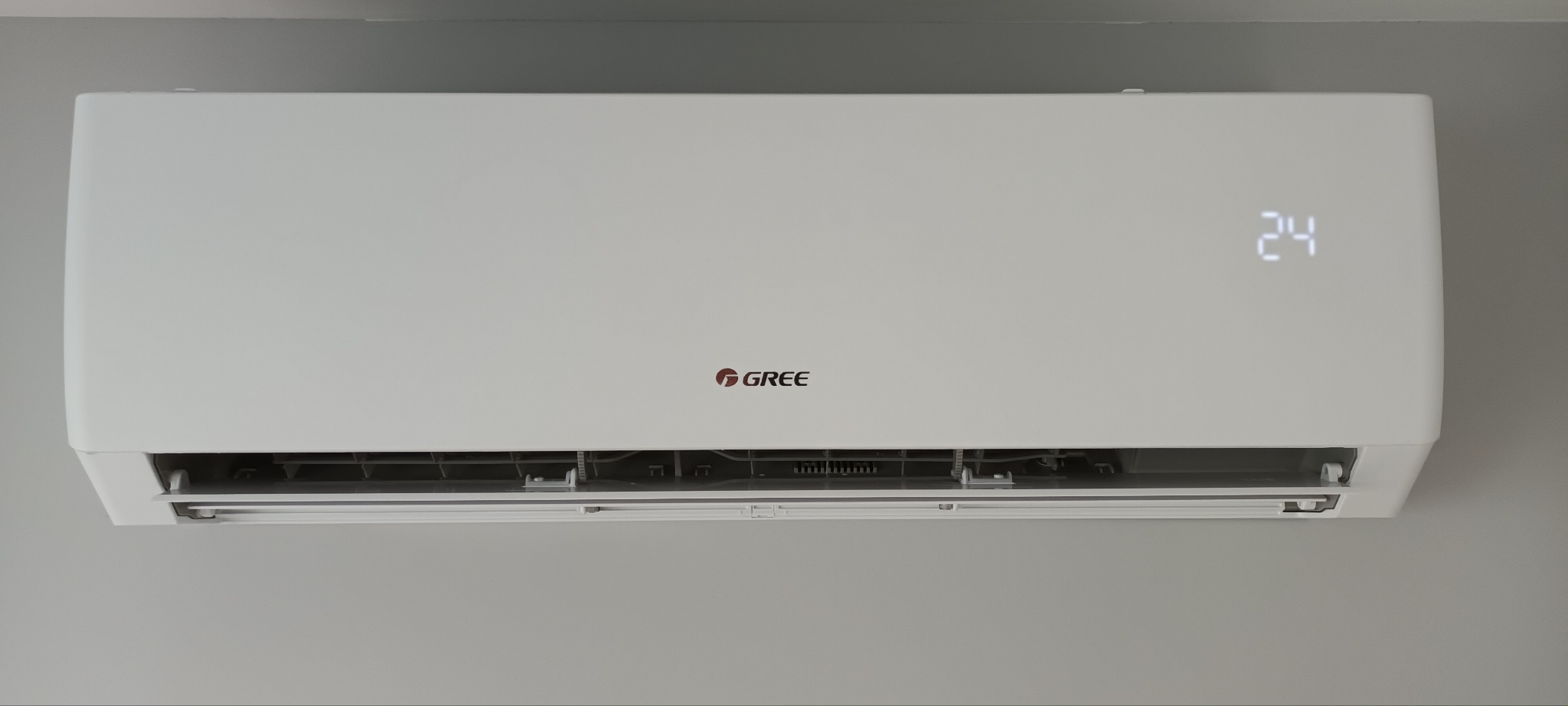 Biały klimatyzator marki Gree zamontowany na szarej ścianie, wyświetlający temperaturę 24 stopnie Celsjusza.