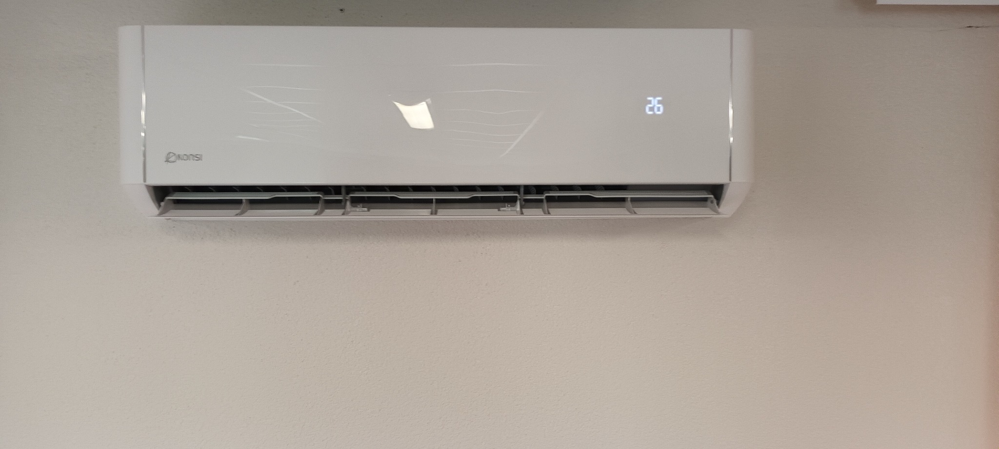 Biała klimatyzacja marki Nonsi zamontowana na kremowej ścianie, wyświetlająca temperaturę 26 stopni Celsjusza.