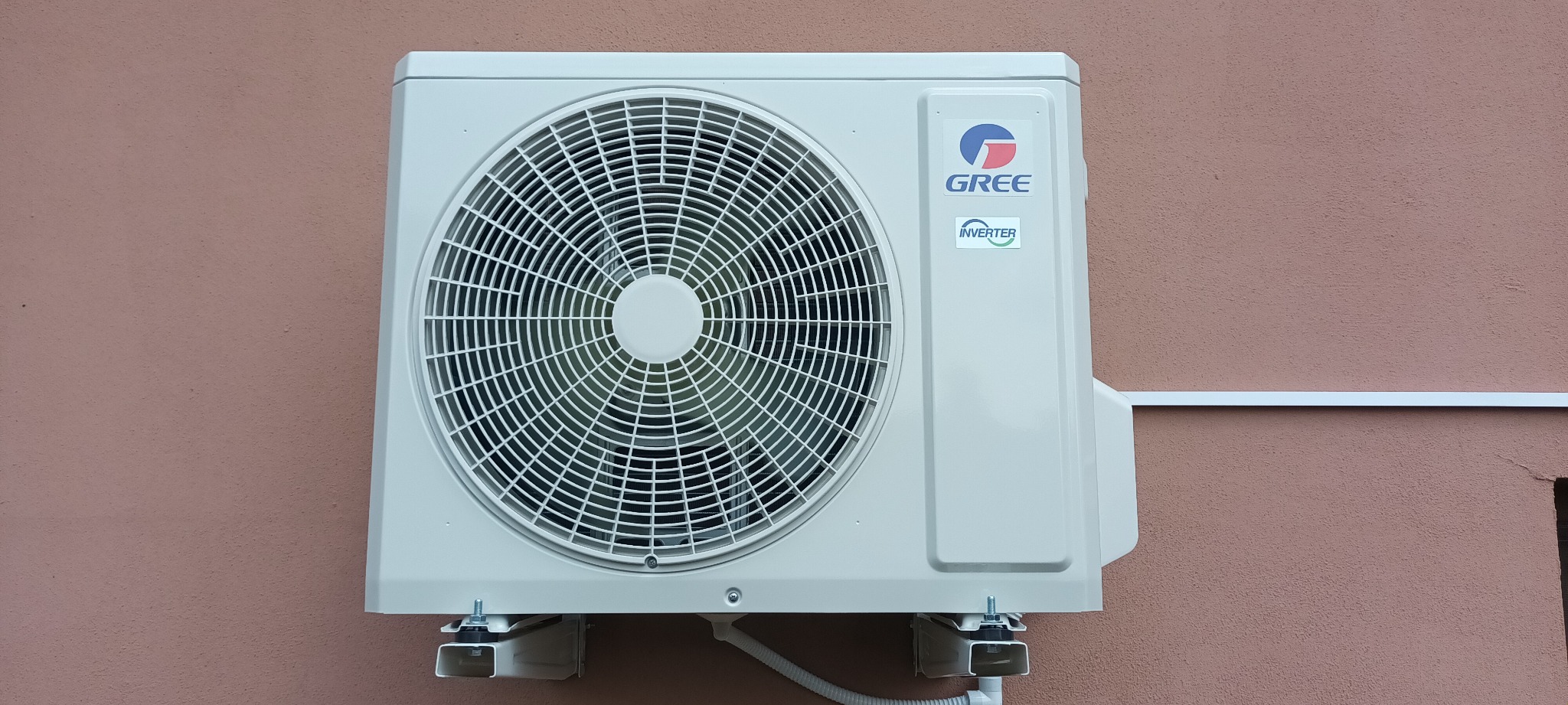 Zewnętrzna jednostka klimatyzacji marki Gree zamontowana na ścianie budynku o różowej elewacji. Widoczne logo i oznaczenie Inverter. Białe przewody i wsporniki montażowe.