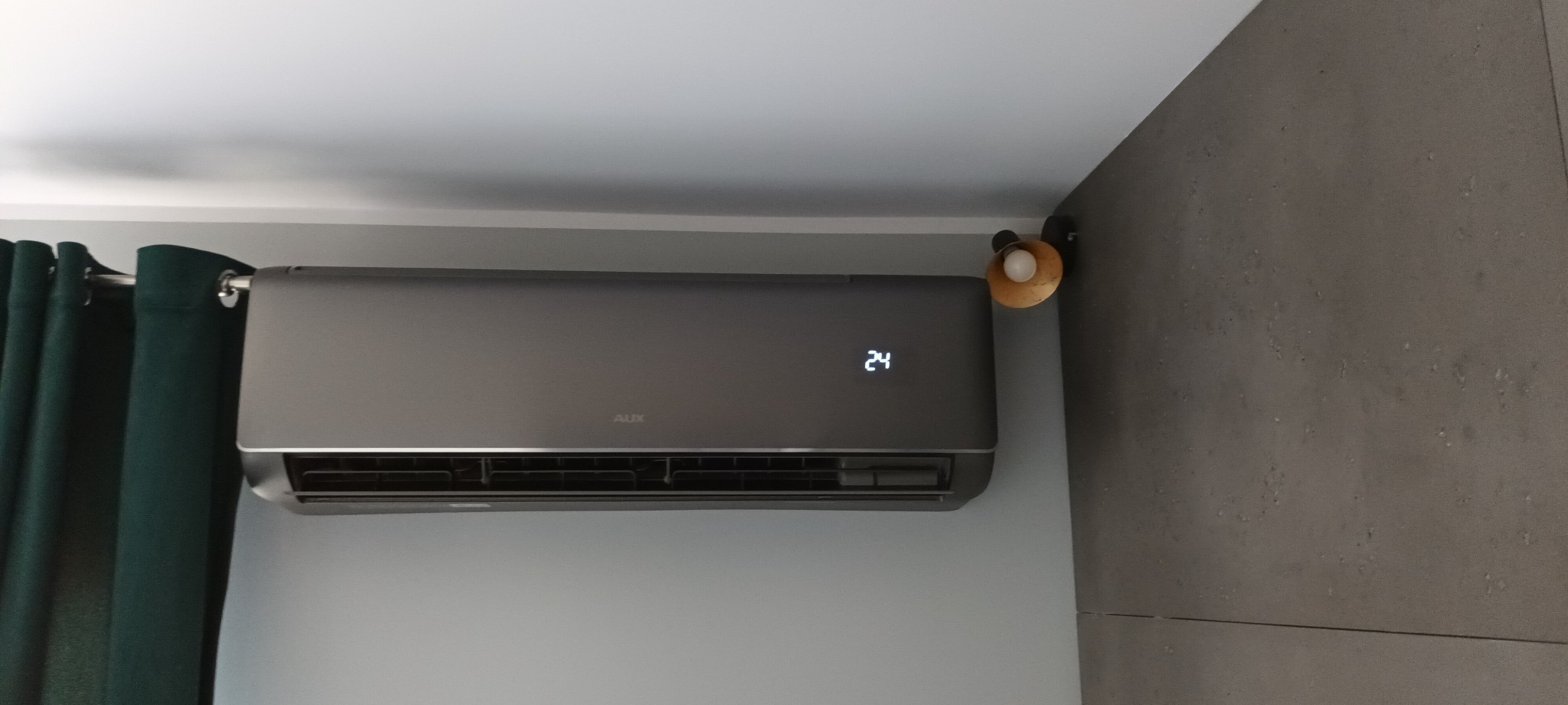 Szary klimatyzator AUX z wyświetloną temperaturą 24 stopnie, zamontowany na jasnej ścianie, obok ciemnozielonych zasłon i szarej ściany z lampką.