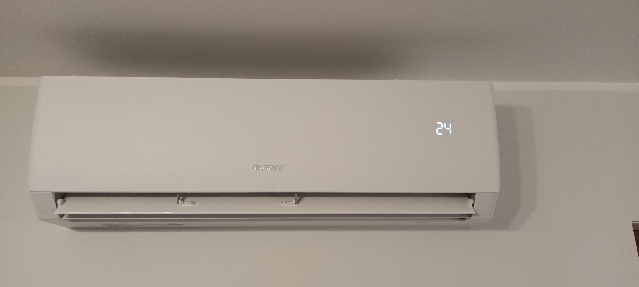 Biały klimatyzator marki Gree zamontowany na jasnej ścianie, wyświetlający temperaturę 24 stopnie Celsjusza.