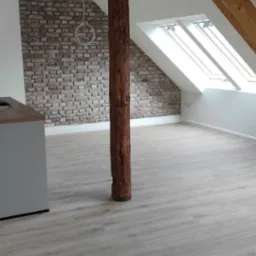 Poddasze po remoncie z drewnianą kolumną pośrodku, wykończone panelami podłogowymi i ścianą z cegły dekoracyjnej, widoczne okna dachowe i odsłonięta konstrukcja drewniana dachu.