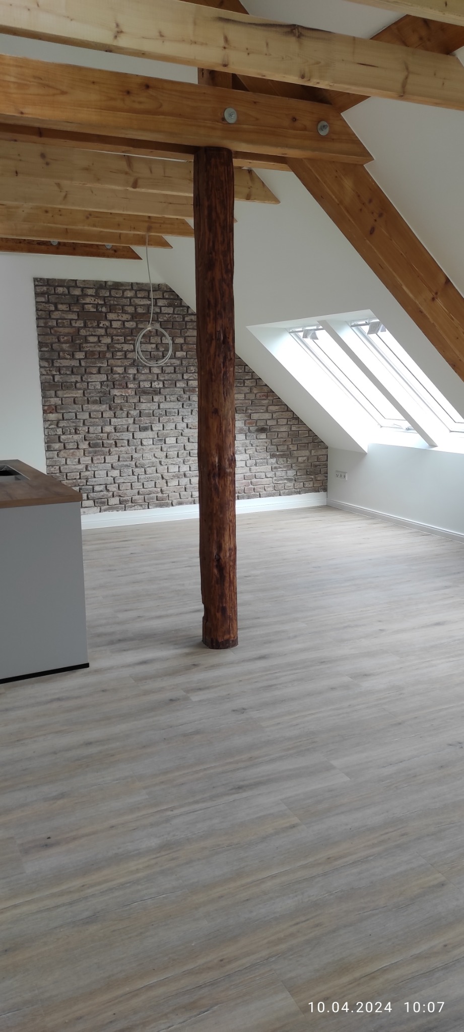 Poddasze po remoncie z drewnianą kolumną pośrodku, wykończone panelami podłogowymi i ścianą z cegły dekoracyjnej, widoczne okna dachowe i odsłonięta konstrukcja drewniana dachu.