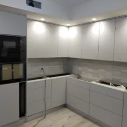 Nowoczesna, jasna kuchnia z białymi, gładkimi frontami szafek bez uchwytów, z zabudowaną czarną kuchenką mikrofalową i piekarnikiem, w trakcie montażu, widoczne podłączenia elektryczne i brak blatu.