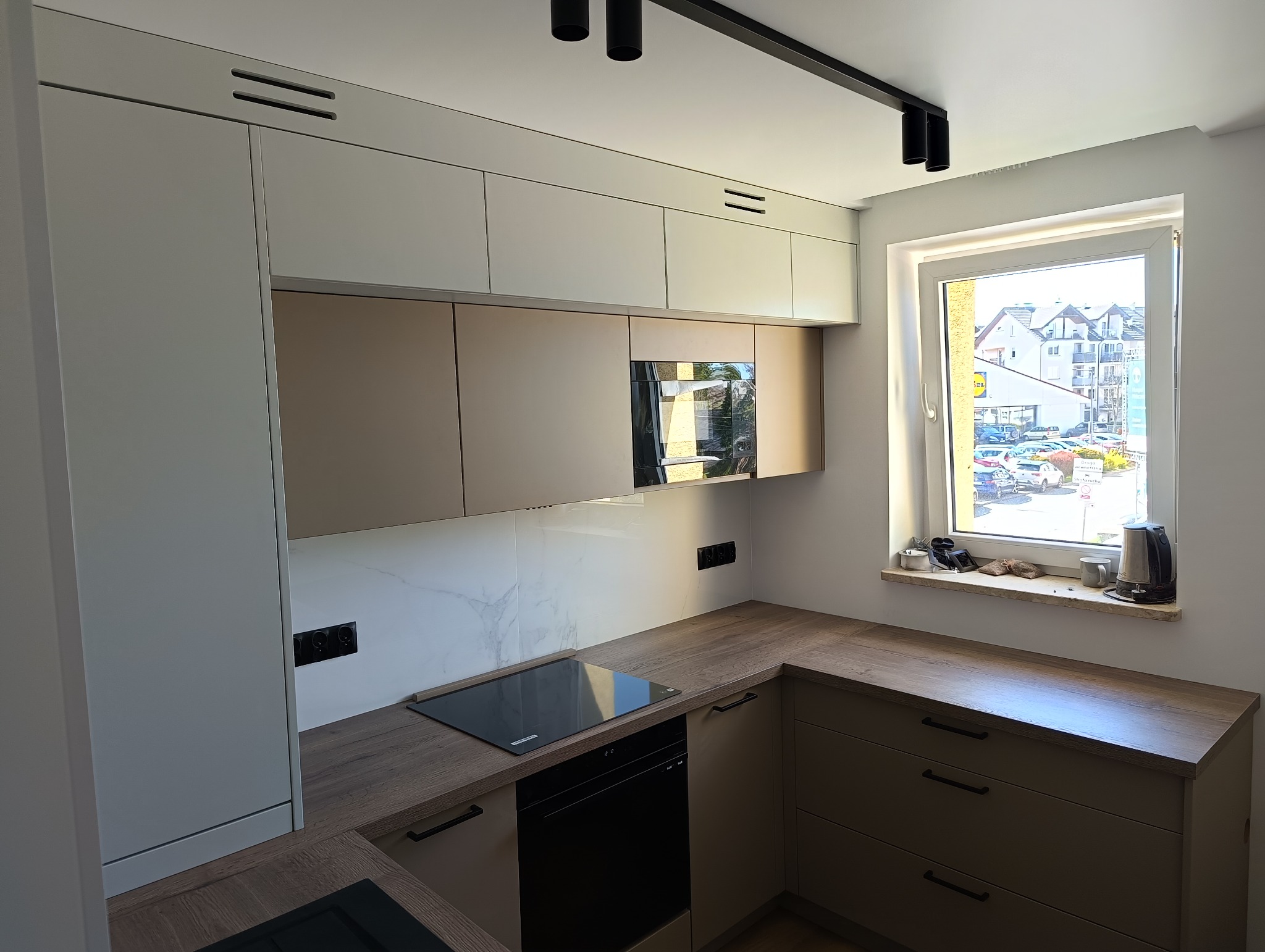 Nowoczesna, jasna kuchnia z drewnianymi blatami i białymi górnymi szafkami, widok na okno z widocznym budynkiem Lidl po drugiej stronie ulicy.