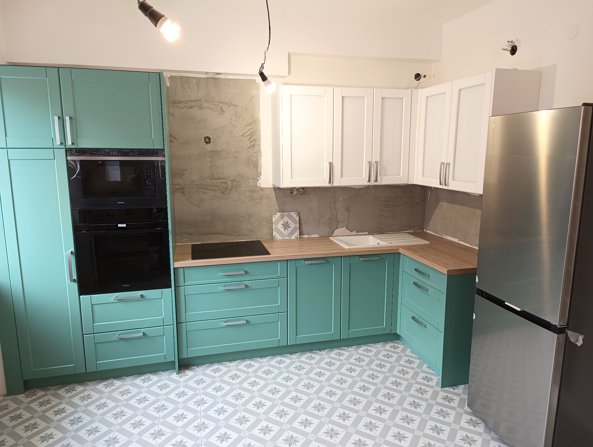 Fragment kuchni z turkusowymi szafkami dolnymi i białymi górnymi, drewnianym blatem, płytą indukcyjną i zlewem. Widoczna również lodówka ze stali nierdzewnej i surowa ściana w tle.