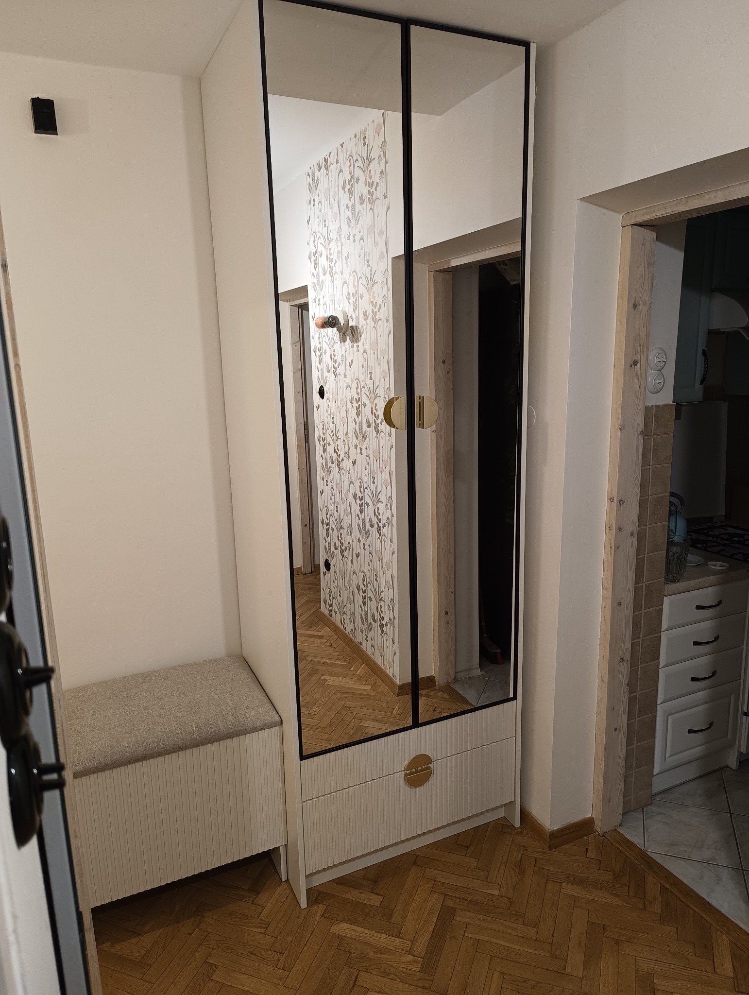 Wysoka szafa z lustrzanymi drzwiami i szufladami w przedpokoju, obok siedzisko z tapicerowanym blatem, widoczne odbicie tapety we wzory.