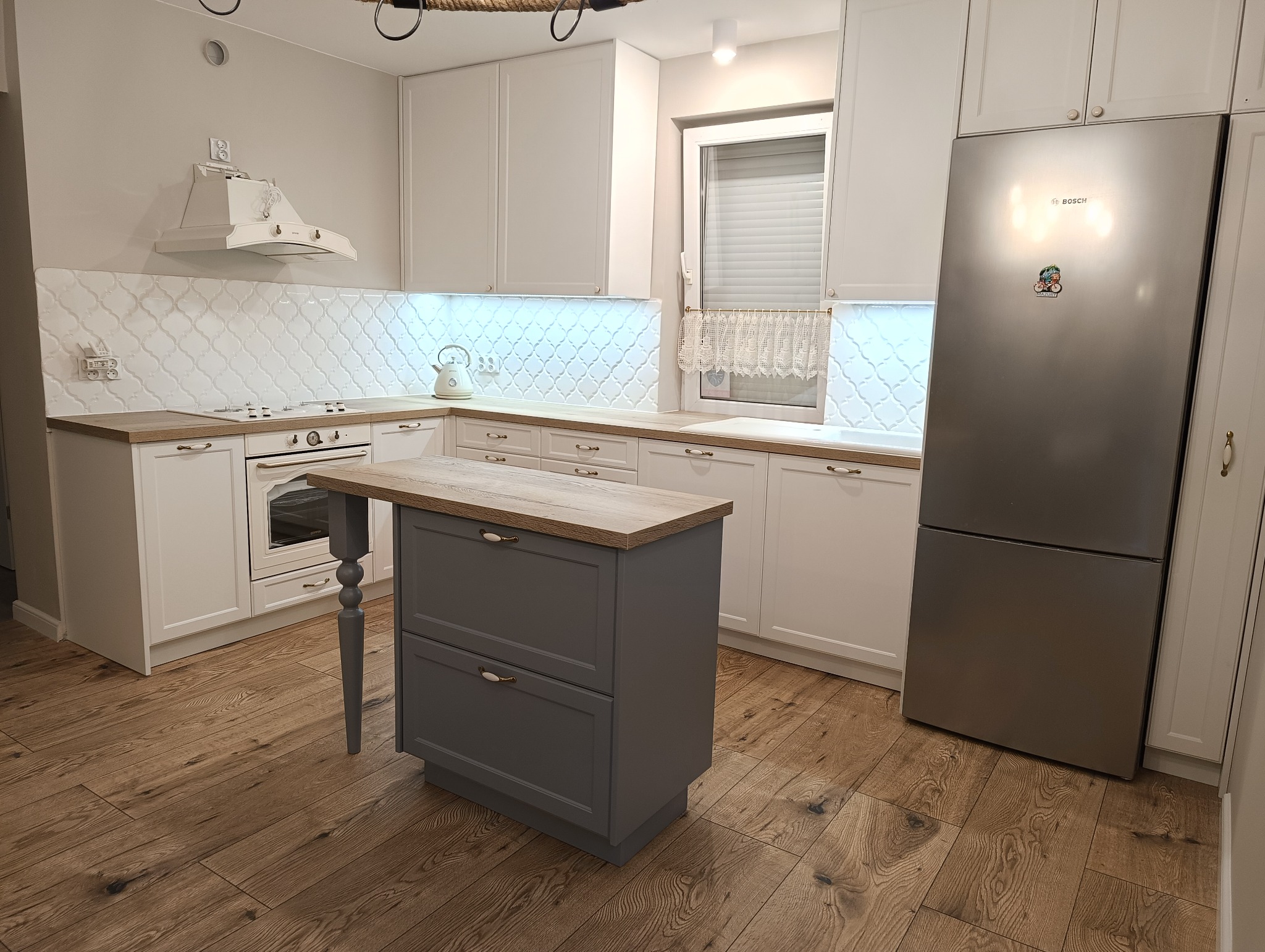 Jasna kuchnia z białymi szafkami i drewnianym blatem, szara wyspa kuchenna z dwoma szufladami, białe płytki na ścianie w marokański wzór, oświetlenie LED pod szafkami, drewniana podłoga.