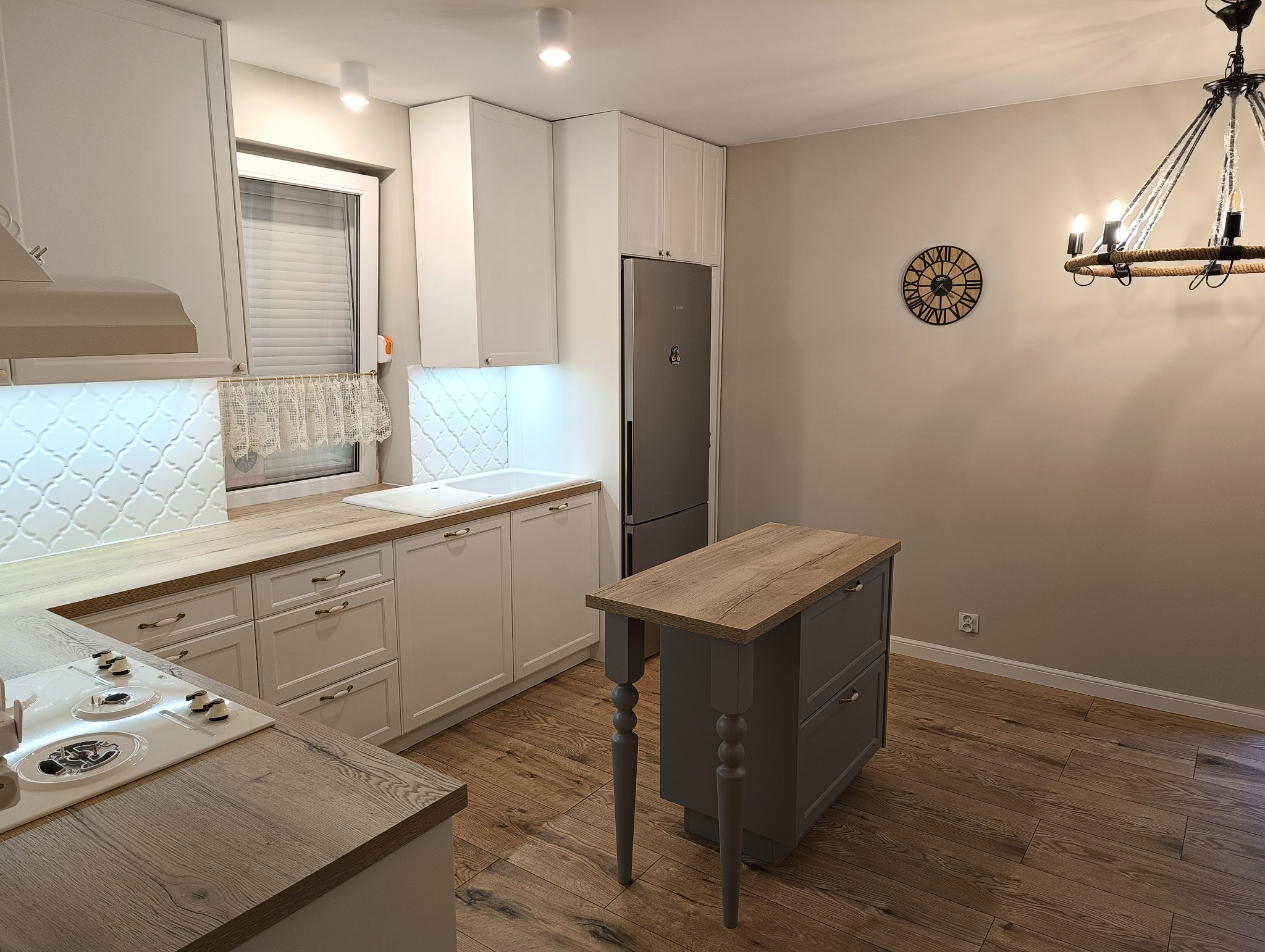 Jasna kuchnia z białymi szafkami, drewnianymi blatami i szarą wyspą na drewnianej podłodze; dekoracyjny zegar na ścianie.