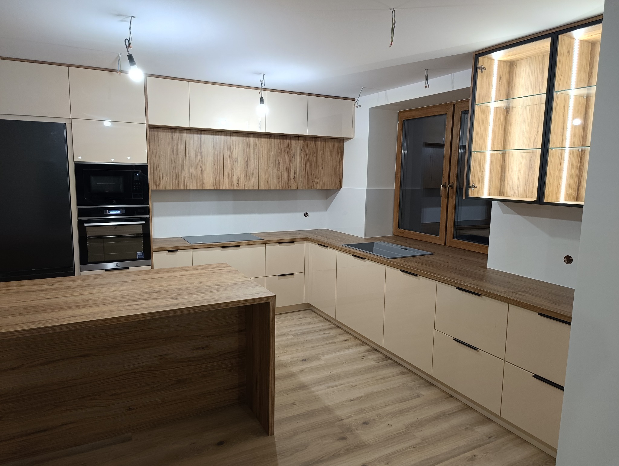 Nowoczesna kuchnia z beżowymi szafkami, drewnianym blatem i wyspą, oświetleniem LED w górnej szafce z przeszklonymi drzwiczkami, oraz niezabezpieczonymi kablami od oświetlenia sufitowego.