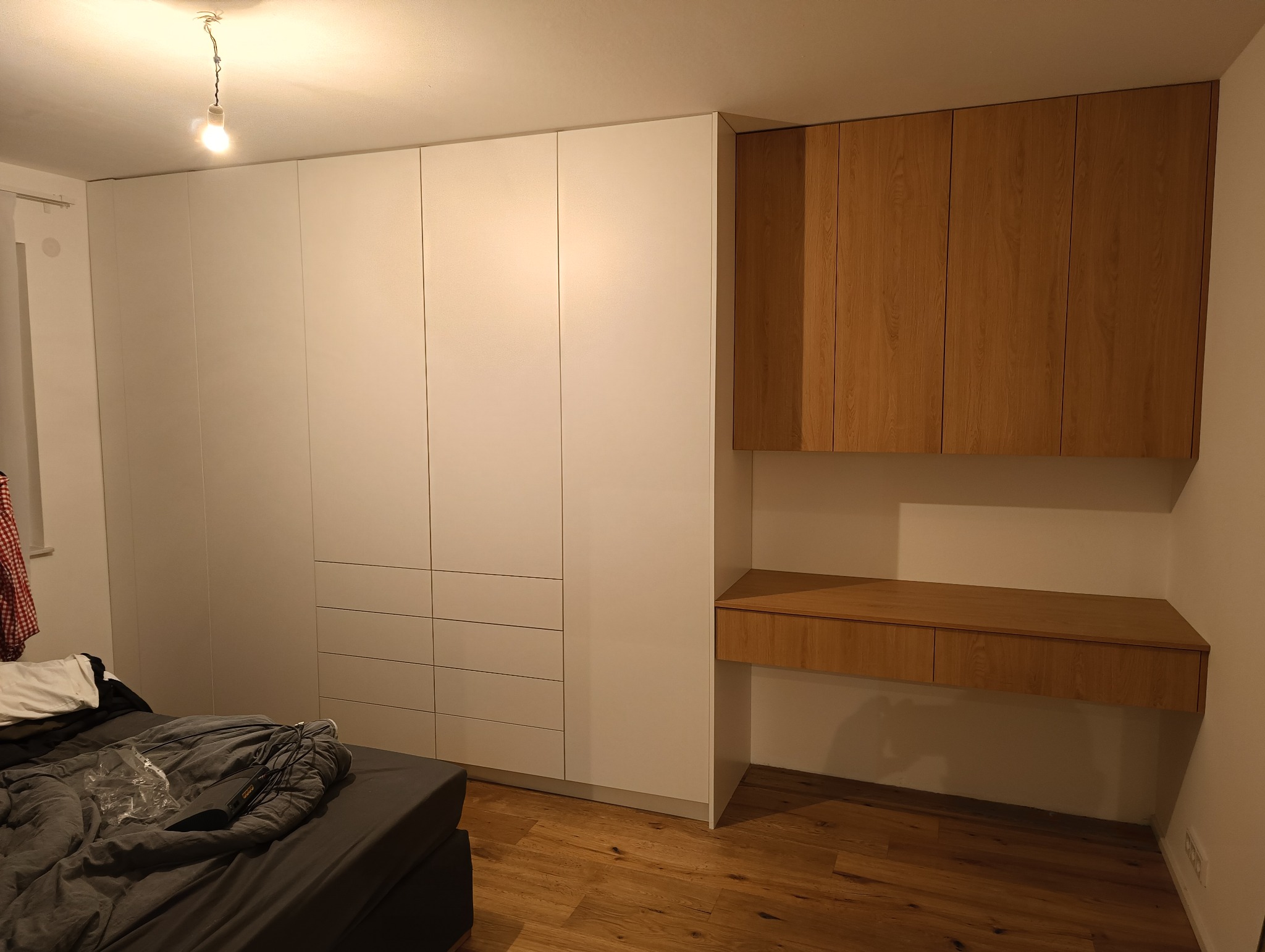 Biała szafa wnękowa z drewnianym biurkiem i szafkami wiszącymi, oświetlona żarówką bez klosza, widoczna część łóżka.