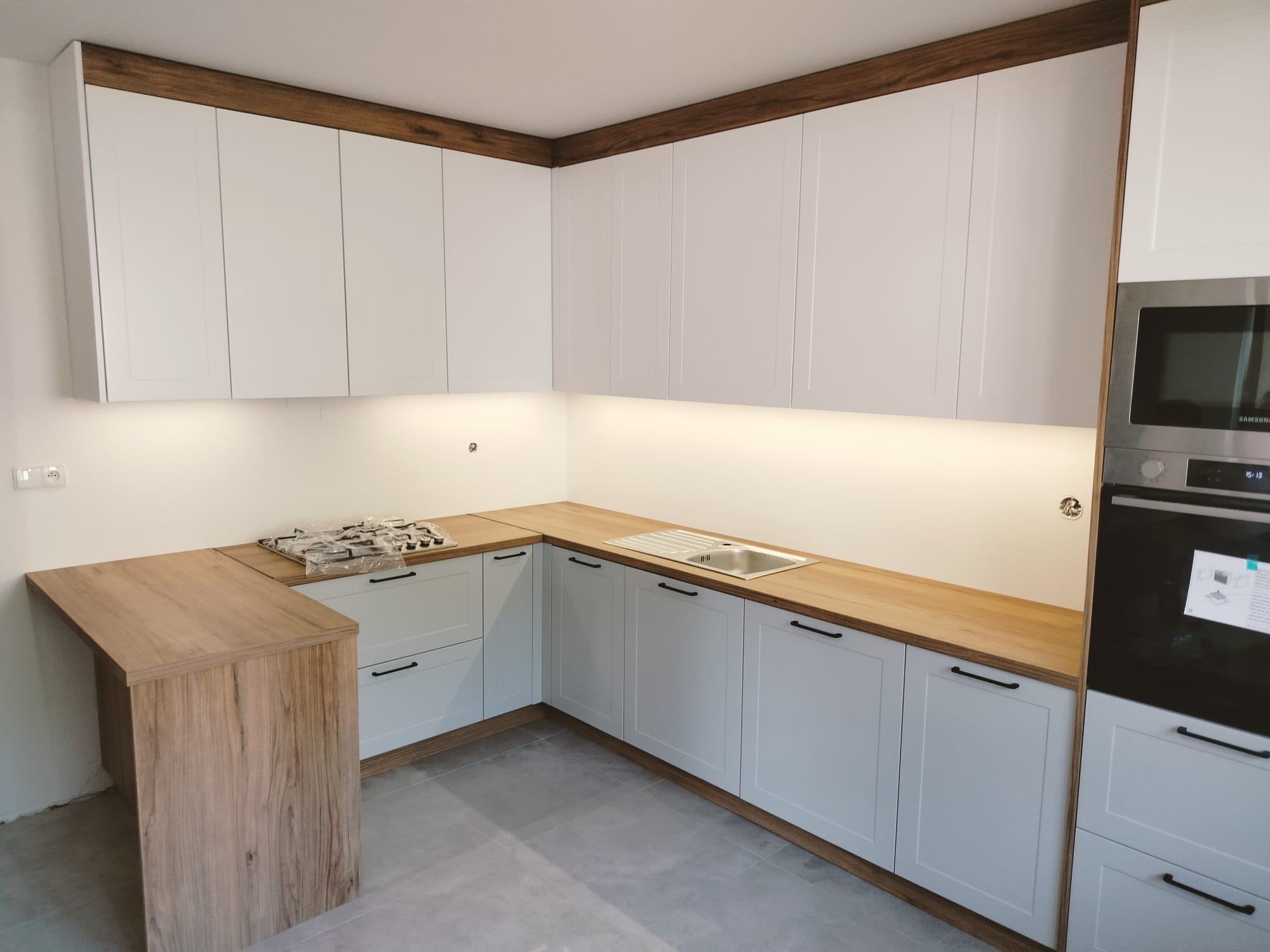 Jasna kuchnia z białymi szafkami i drewnianym blatem, widoczny zlew, płyta gazowa w folii i zabudowany piekarnik z mikrofalówką.