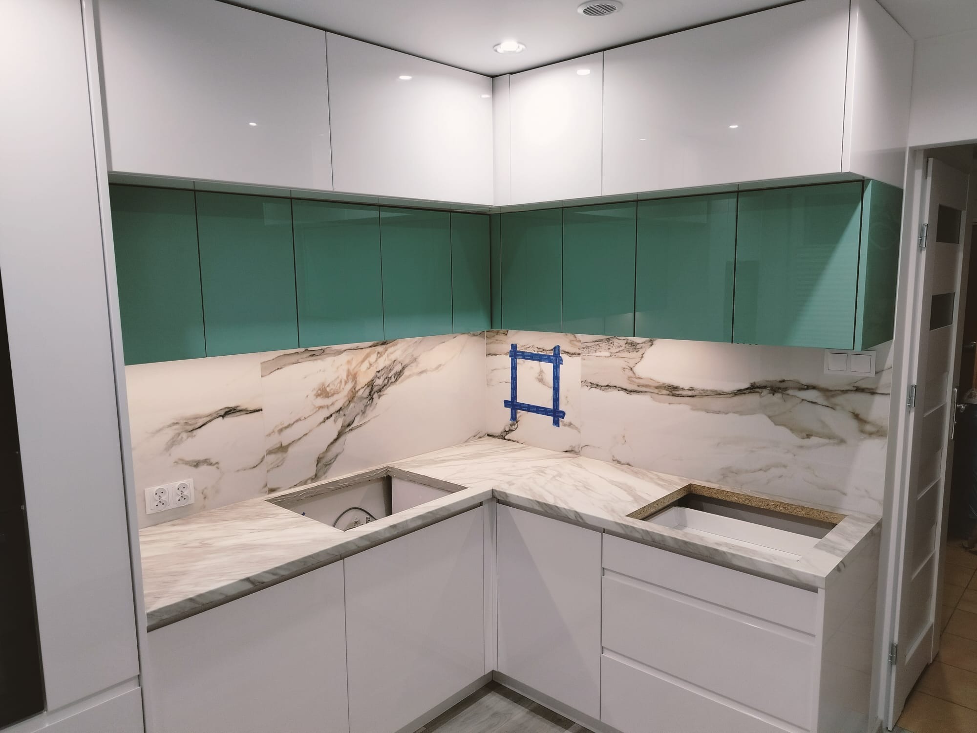 Wnętrze kuchni w Przytkowicach w trakcie montażu: białe szafki dolne i górne, turkusowe szafki wiszące, marmurowy blat z wyciętymi otworami na zlew, na ścianie fragment płytki marmurowej...