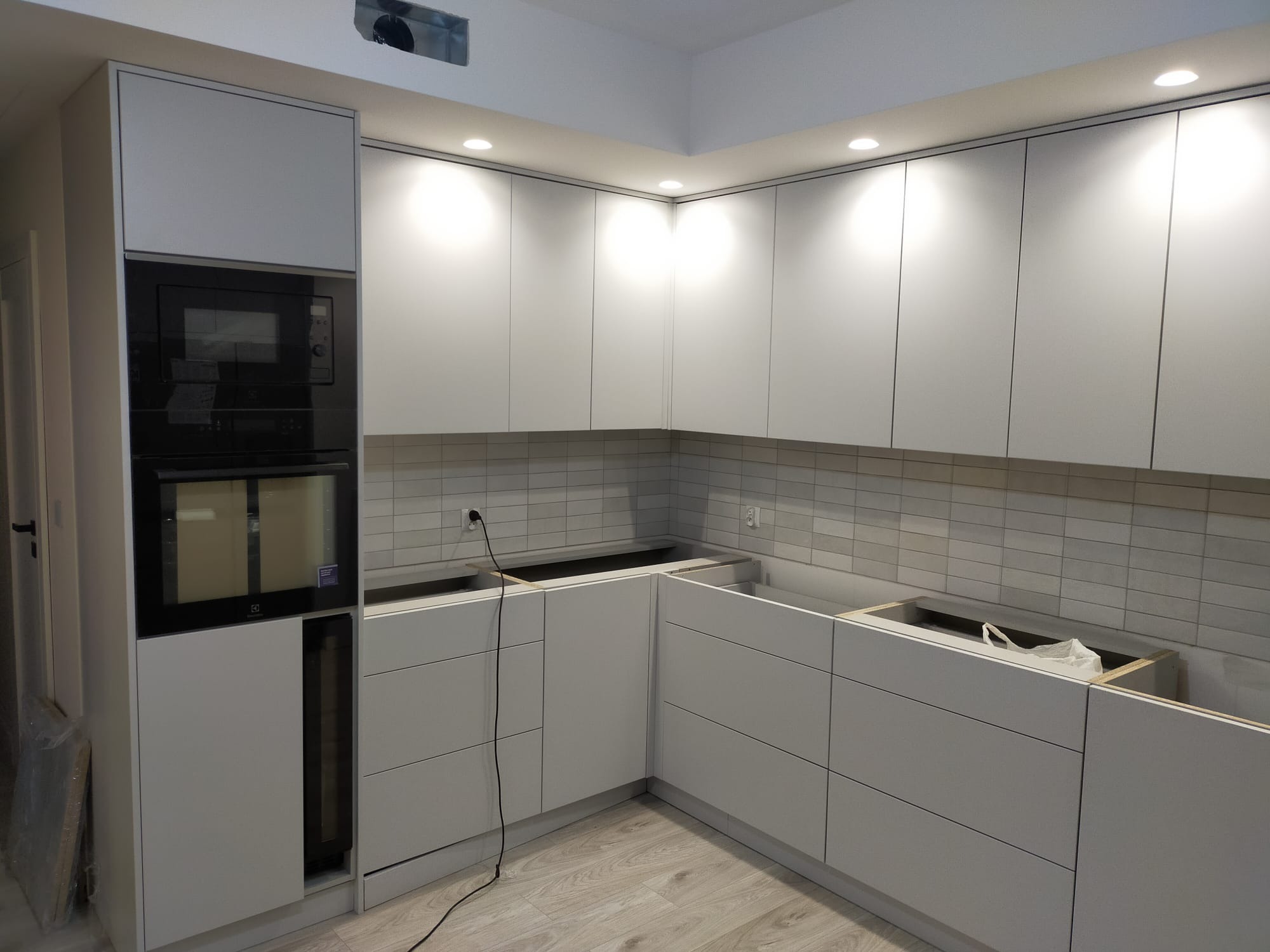 Nowoczesna, jasna kuchnia z białymi, gładkimi frontami szafek bez uchwytów, z zabudowaną czarną kuchenką mikrofalową i piekarnikiem, w trakcie montażu, widoczne podłączenia elektryczne i brak blatu.
