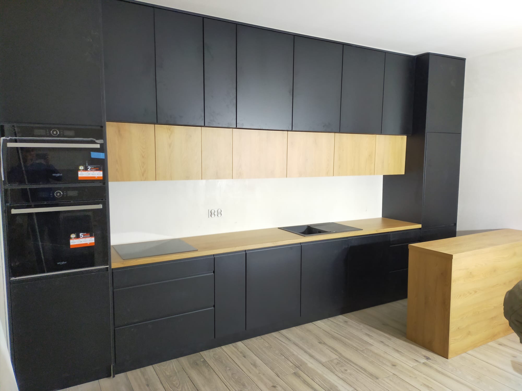 Nowoczesna kuchnia z czarnymi, matowymi frontami szafek i drewnianymi akcentami na blacie i górnych szafkach, z zabudowanymi piekarnikami i płytą grzewczą.