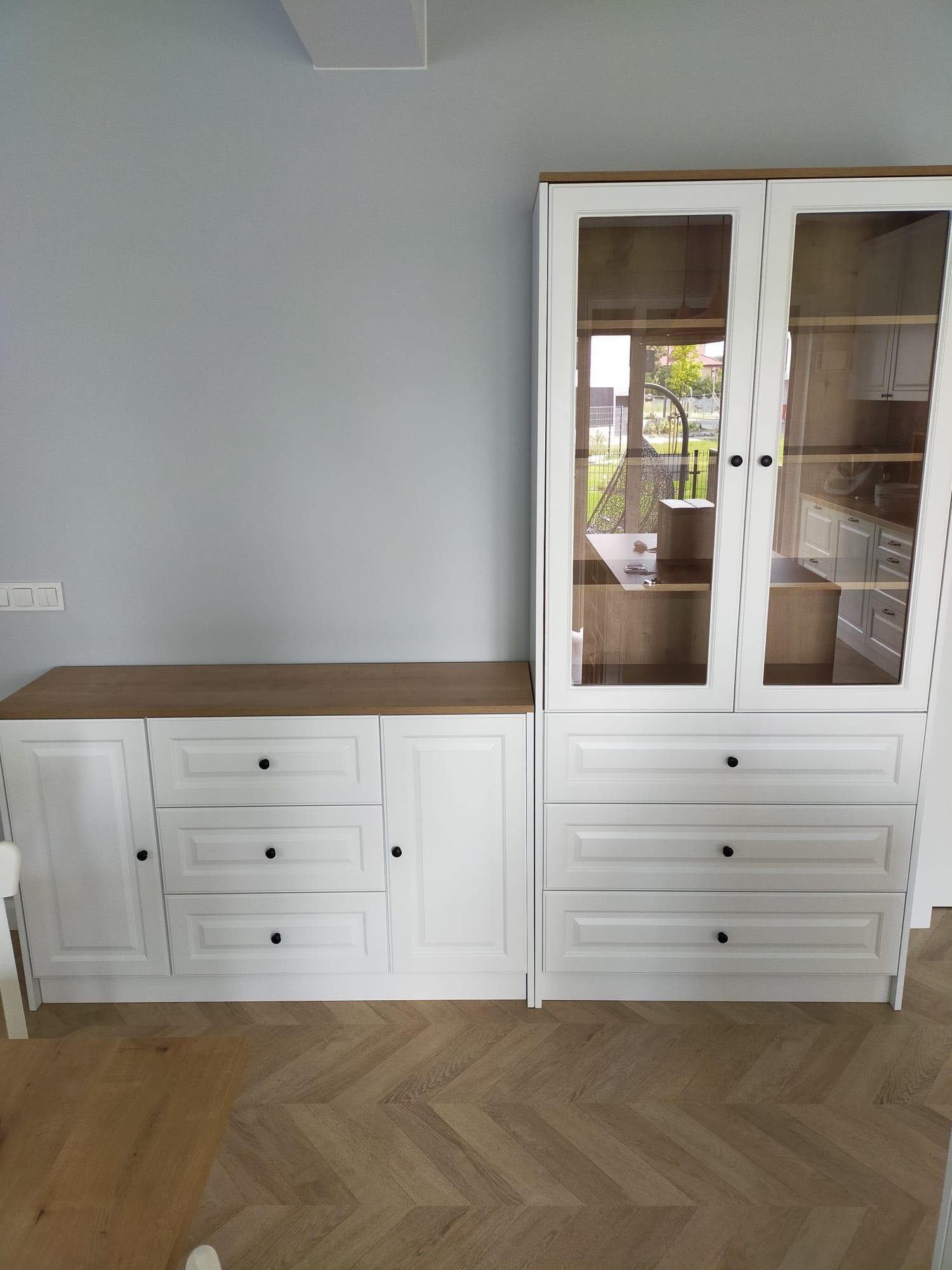 Biała komoda z szufladami i przeszklona witryna z drewnianym blatem na parkiecie w jodełkę, widok na ogród przez szklane drzwi witryny.