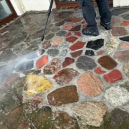 Czyszczenie kolorowej kostki brukowej z kamienia polnego za pomocą myjki ciśnieniowej, widoczne stopy osoby obsługującej urządzenie.