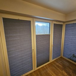 ROLETY -MaAn DOMINIKA SCHÜTZ - Szare, plisowane osłony okienne zamontowane w trzech oknach, z widokiem na zewnątrz i parkietem na podłodze.