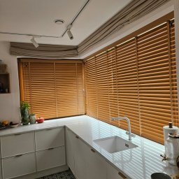 ROLETY -MaAn DOMINIKA SCHÜTZ - Drewniane żaluzje poziome zamontowane w oknach kuchni z białym blatem i zlewozmywakiem, widoczne także lampy sufitowe i zasłony rzymskie nad oknami.